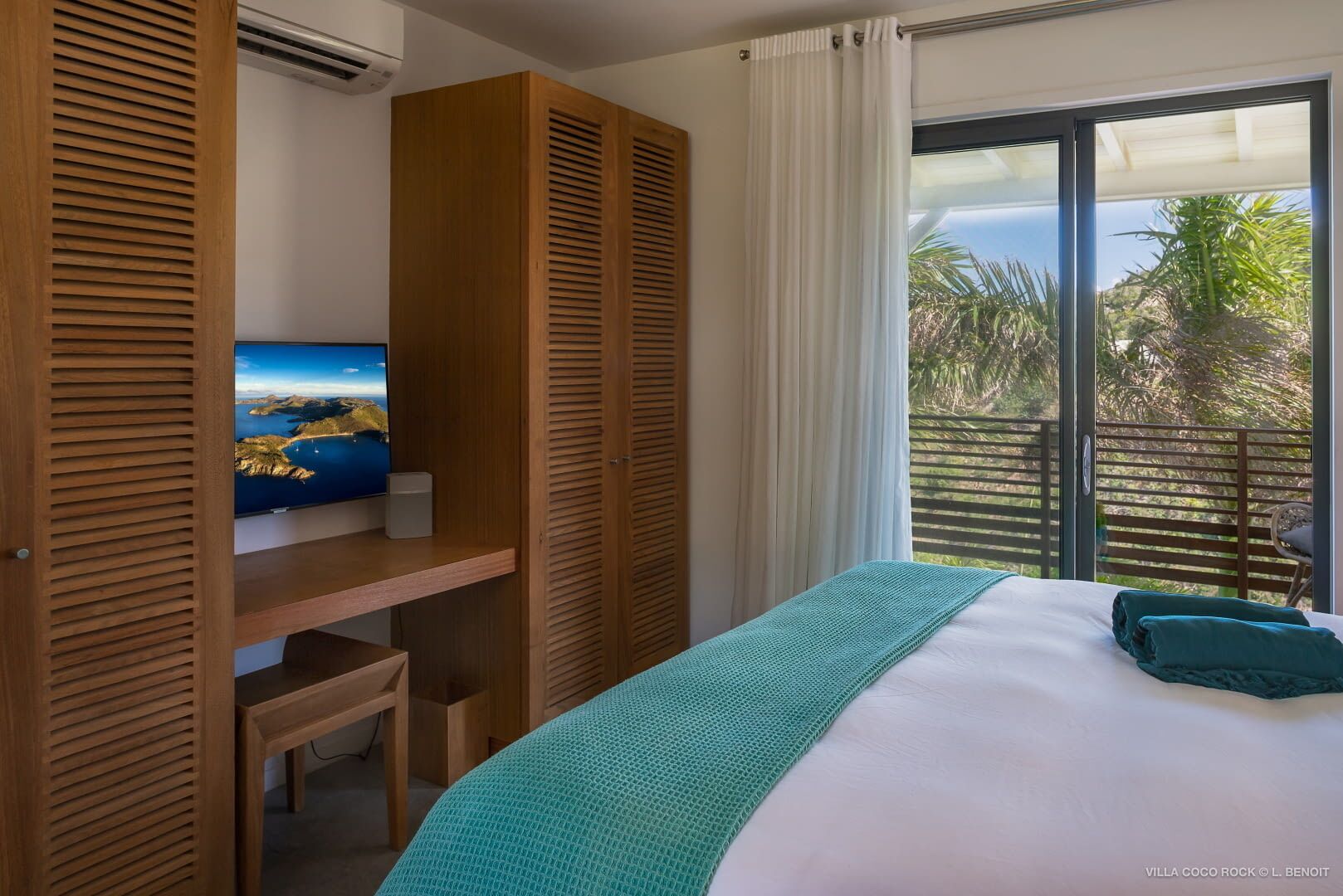 Ein Schlafzimmer mit Bett, einem Fernseher auf einer Holzwand, einem Balkon mit Blick auf Palmen und einem Fenster mit weißen Vorhängen.
