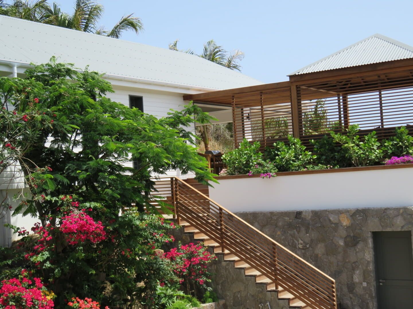 Una casa moderna con una escalera de madera que lleva a un espacio exterior cubierto, rodeada de exuberante vegetación y plantas florecientes.
