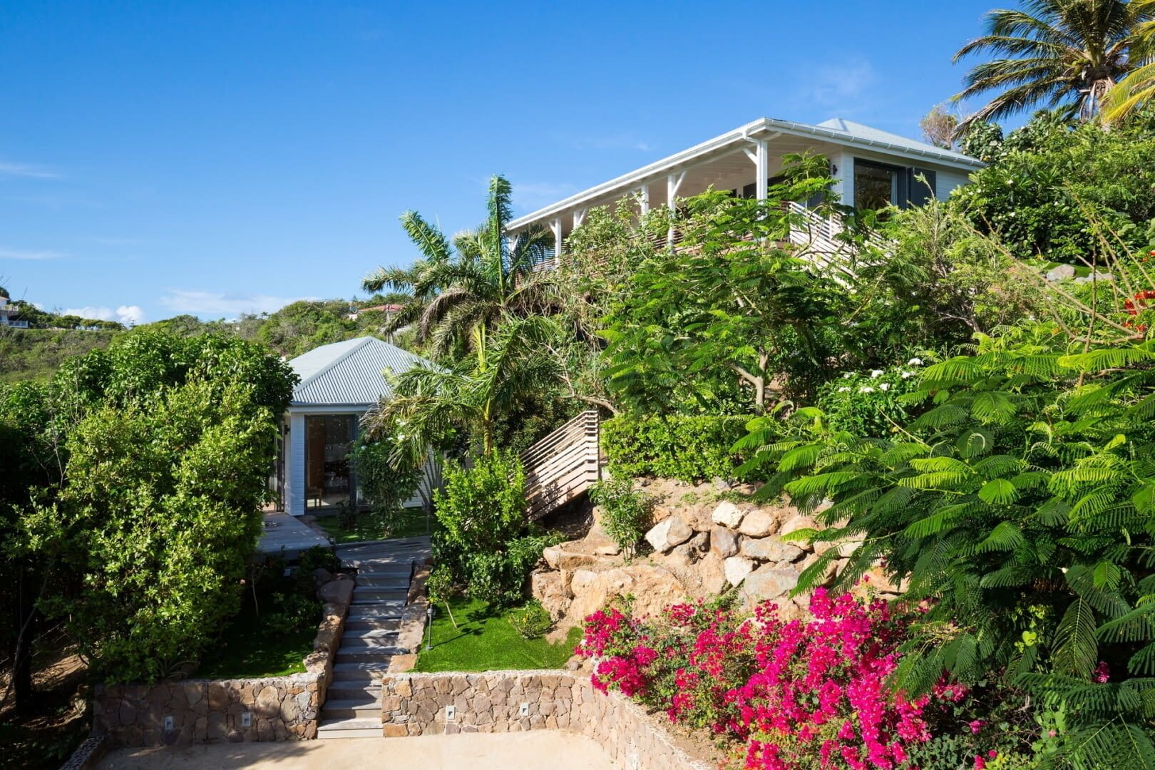Una casa con una escalera de piedra que conduce a un porche cubierto, rodeada de abundante vegetación y plantas en flor.