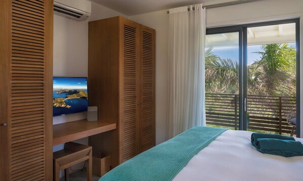A bedroom with a bed, a TV on a wooden shelf, a balcony with a view of palm trees, and a window with white curtains.