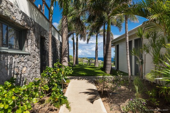 A pathway leading to a building with a stone facade, surrounded by palm trees and a view of the ocean.