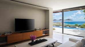 Ein modernes Wohnzimmer mit großem Flachbildfernseher, einer Holz-Unterhaltungsecke und einem Couchtisch mit einer Vase. Eine große Schiebeglasdoor führt zu einem Außenbereich mit Pool und Sonnenschirmen.