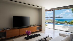 Ein modernes Wohnzimmer mit großem Flachbildfernseher, einer Holz-Unterhaltungsecke und einem Couchtisch mit einer Vase. Eine große Schiebeglasdoor führt zu einem Außenbereich mit Pool und Sonnenschirmen.