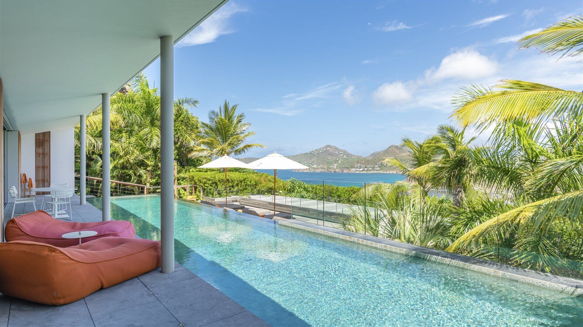 A poolside area with a view of the sea and mountains, a covered seating area, and a clear blue pool.