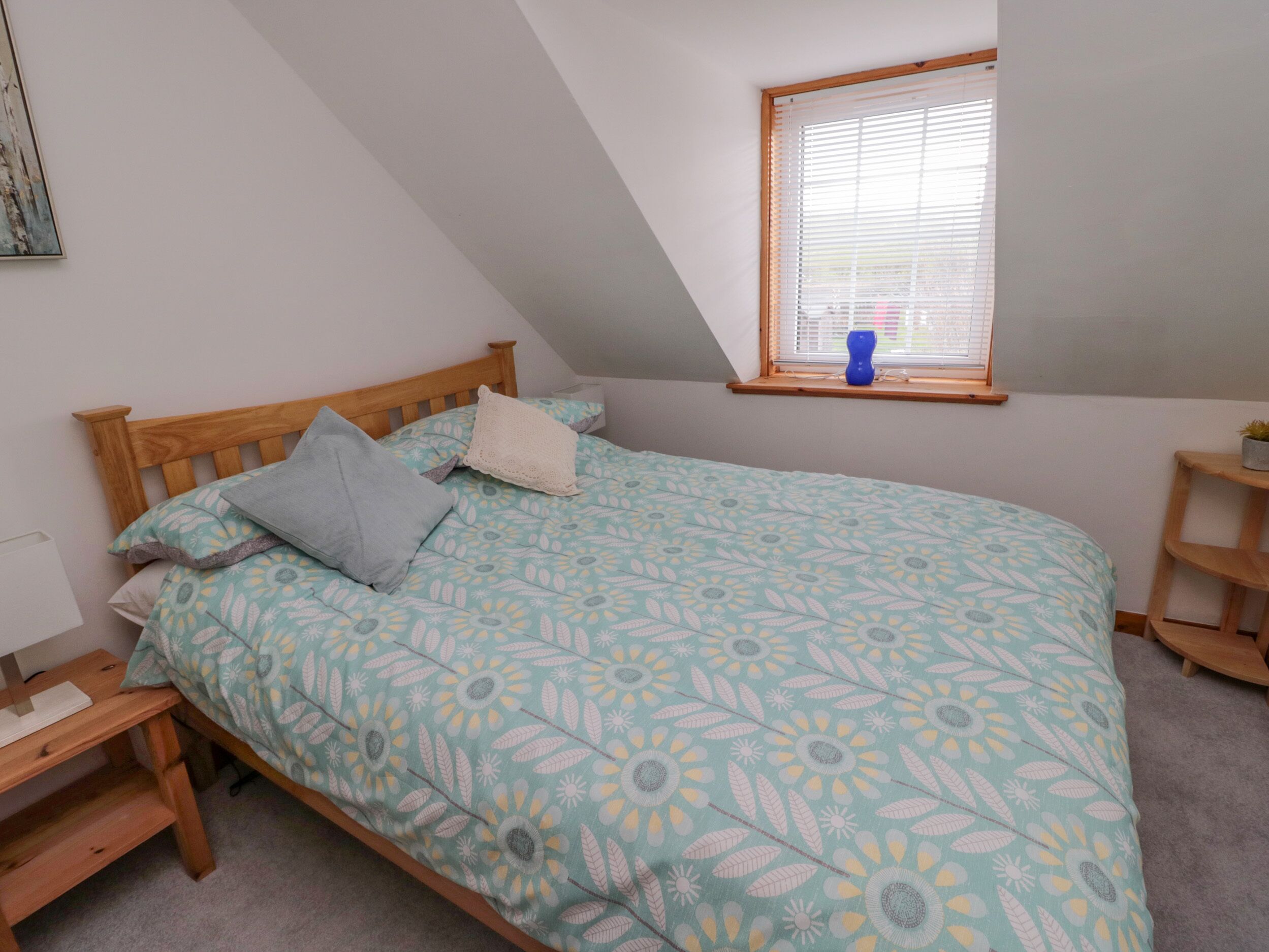 Un dormitorio con techo inclinado, una cama con cubrecama floral, una mesita de noche de madera, una ventana con persianas y un pequeño estante.