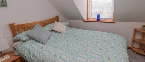 Un dormitorio con techo inclinado, una cama con cubrecama floral, una mesita de noche de madera, una ventana con persianas y un pequeño estante.