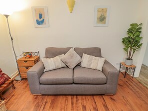 A grey sofa with three cushions, a wooden floor, a lamp, two framed artworks, a potted plant, and a small table with a vase. - 5 Swinton Mill Farm Cottage (Coldstream)