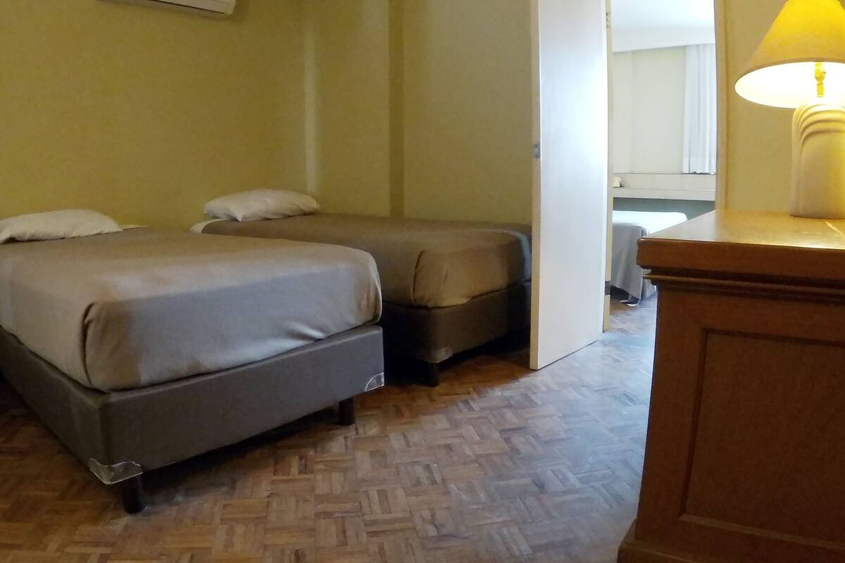 Una habitación con dos camas, piso de madera, un escritorio con lámpara y un espejo.