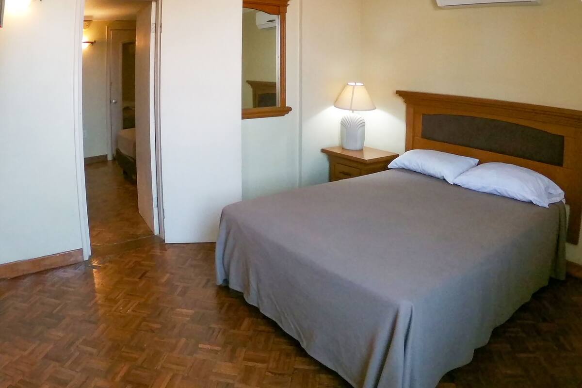 Un dormitorio con piso de madera, una cama con cubrecama gris, una mesita de noche con lámpara y un espejo en la pared.