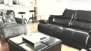 A living room with a black leather sofa, a matching ottoman, and a coffee table. There is a kitchen in the background with a refrigerator, stove, and bar stools.