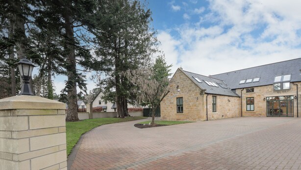 A stone building with a brick driveway, large trees, and a clear sky.