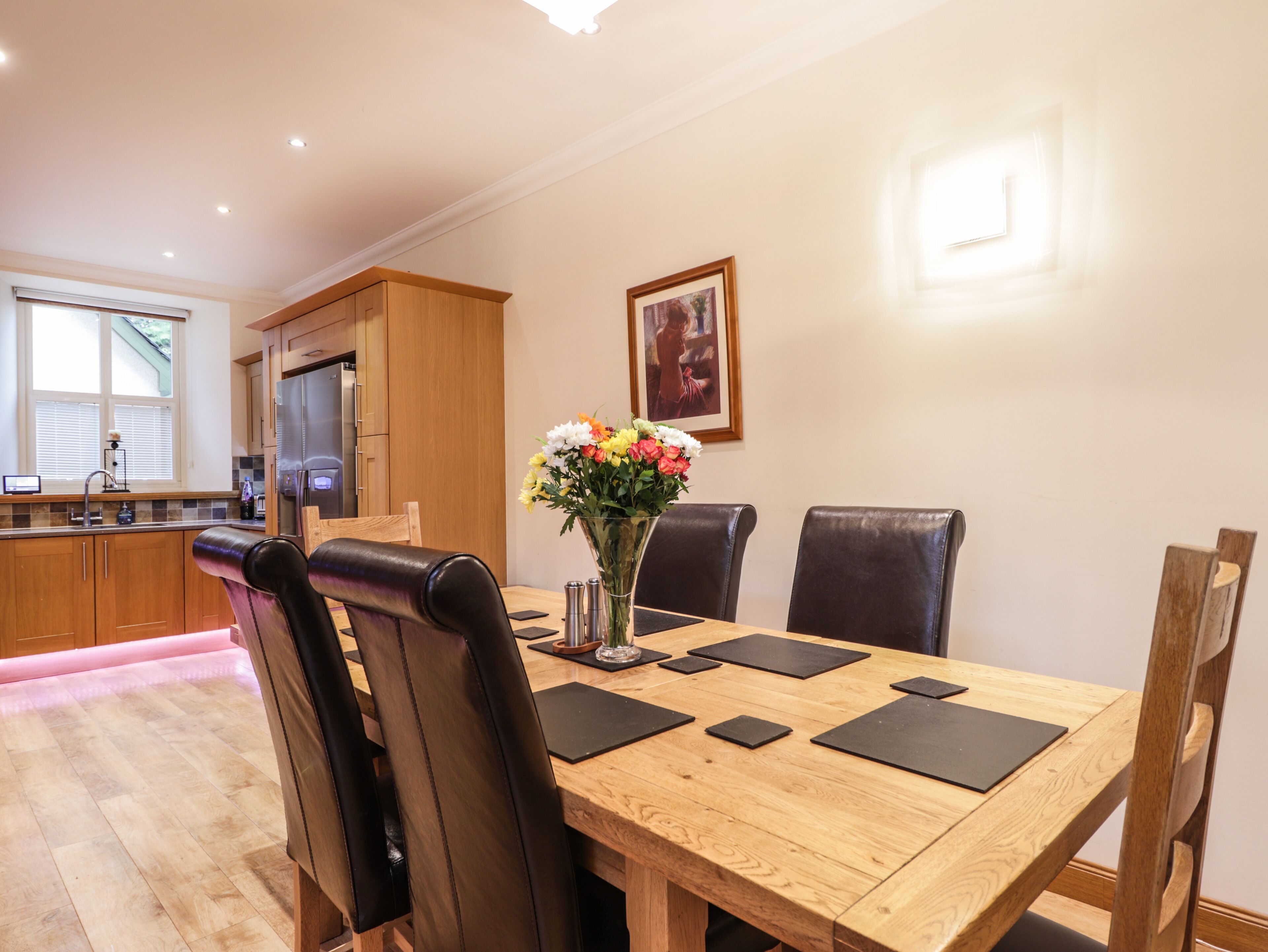 A dining area with a wooden table, black chairs, and a vase of flowers. There is a kitchen in the background with wooden cabinets and a refrigerator.