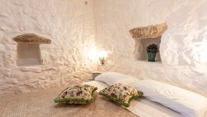 Quarto com paredes brancas, cama com almofadas brancas e florais, uma pequena planta verde e um luminário fixado na parede.