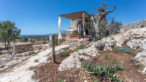 Uma casa de pedra com pérgola de madeira, rodeada por terreno rochoso e vegetação rasteira.