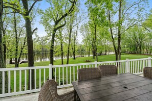 Une terrasse avec une table et des chaises donnant sur un lac, entourée d’arbres et d’une clôture blanche.