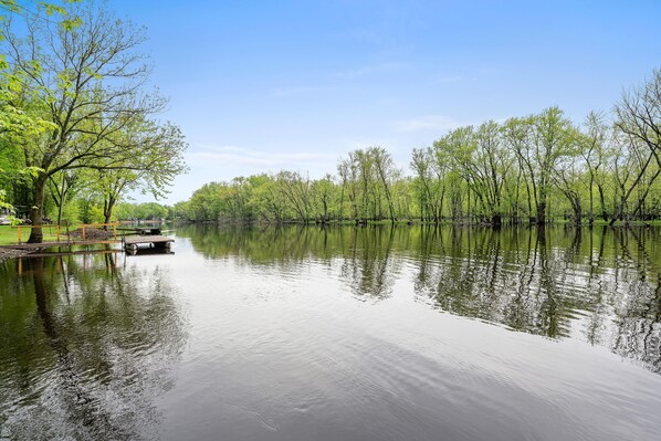 Une rivière paisible, bordée d’arbres dont le reflet se mire dans l’eau, une jetée en bois et un ciel dégagé.
