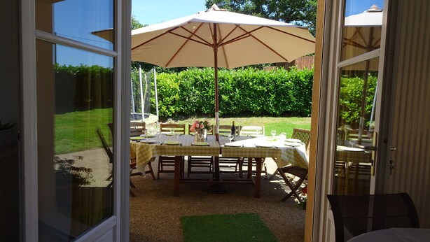 An open sliding glass door leading to an outdoor patio area with a table set for dining, an umbrella, and a view of a well-maintained garden. - FAMILY HOME, former farmhouse with exceptional views. (Pommerieux)