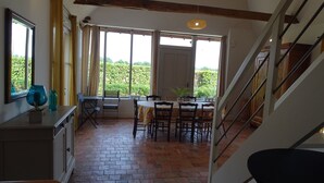 A dining area with a brick floor, wooden chairs, a table with a tablecloth, and a view of greenery through large windows. - FAMILY HOME, former farmhouse with exceptional views. (Pommerieux)
