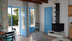 En stue med fjernsyn, sofa, spisebord med stole og en balkon med mønstrede gardiner.