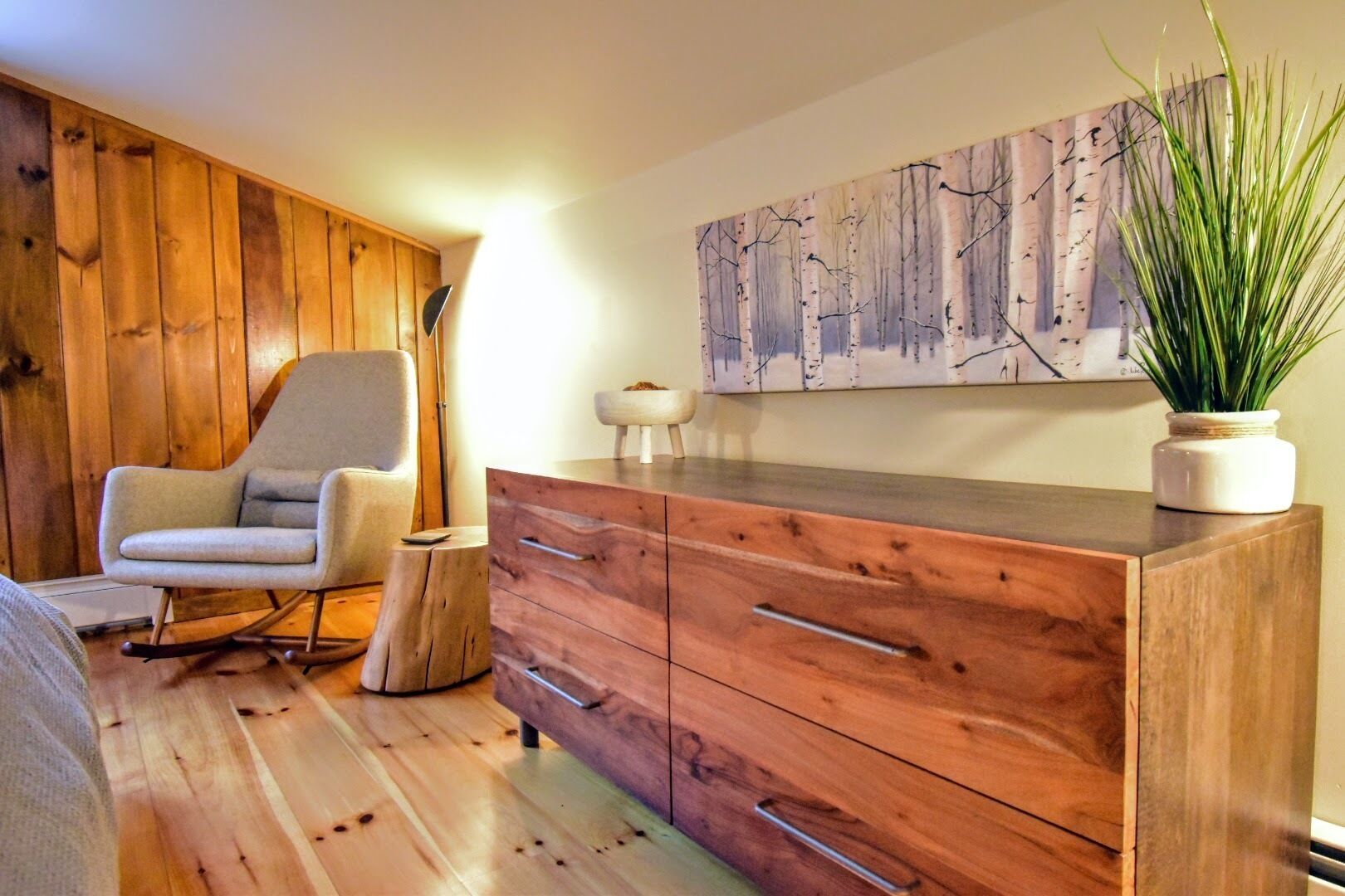 A room with a wooden floor, a rocking chair, a wooden dresser with drawers, a wall-mounted painting, and a potted plant.