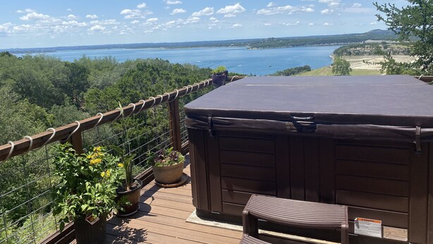 Eine Holzterrasse mit Whirlpool, Topfpflanzen und Blick auf einen See und ferne Hügel.