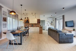 Une cuisine moderne dotée de luminaires suspendus, un coin repas avec une table et des chaises en bois, et un salon avec un canapé gris et un téléviseur à écran plat.