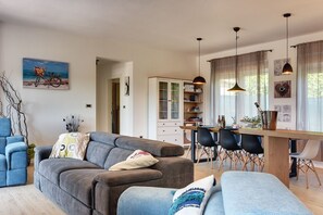 Un salon moderne avec un canapé gris, un fauteuil bleu et une table à manger en bois avec des chaises. On y trouve également une bibliothèque et un tableau accroché au mur.