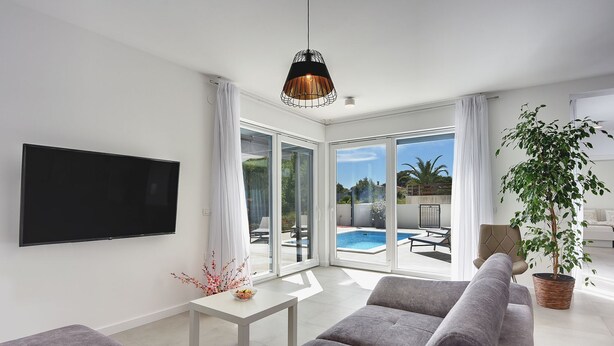 A modern living room with a flat-screen TV, grey sofas, a coffee table with flowers, a hanging pendant light, and a view of a swimming pool and outdoor seating area through large glass doors. - Modern villa with private pool, sun terrace only 2 km from the beach (Medulin)