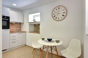 Una cocina moderna con armarios blancos, horno integrado y una mesa redonda con sillas. Un reloj de pared grande está colgado sobre la mesa.