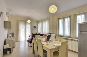 A dining area with a wooden table, chairs, and a chandelier. There is a sofa and a television in the background.
