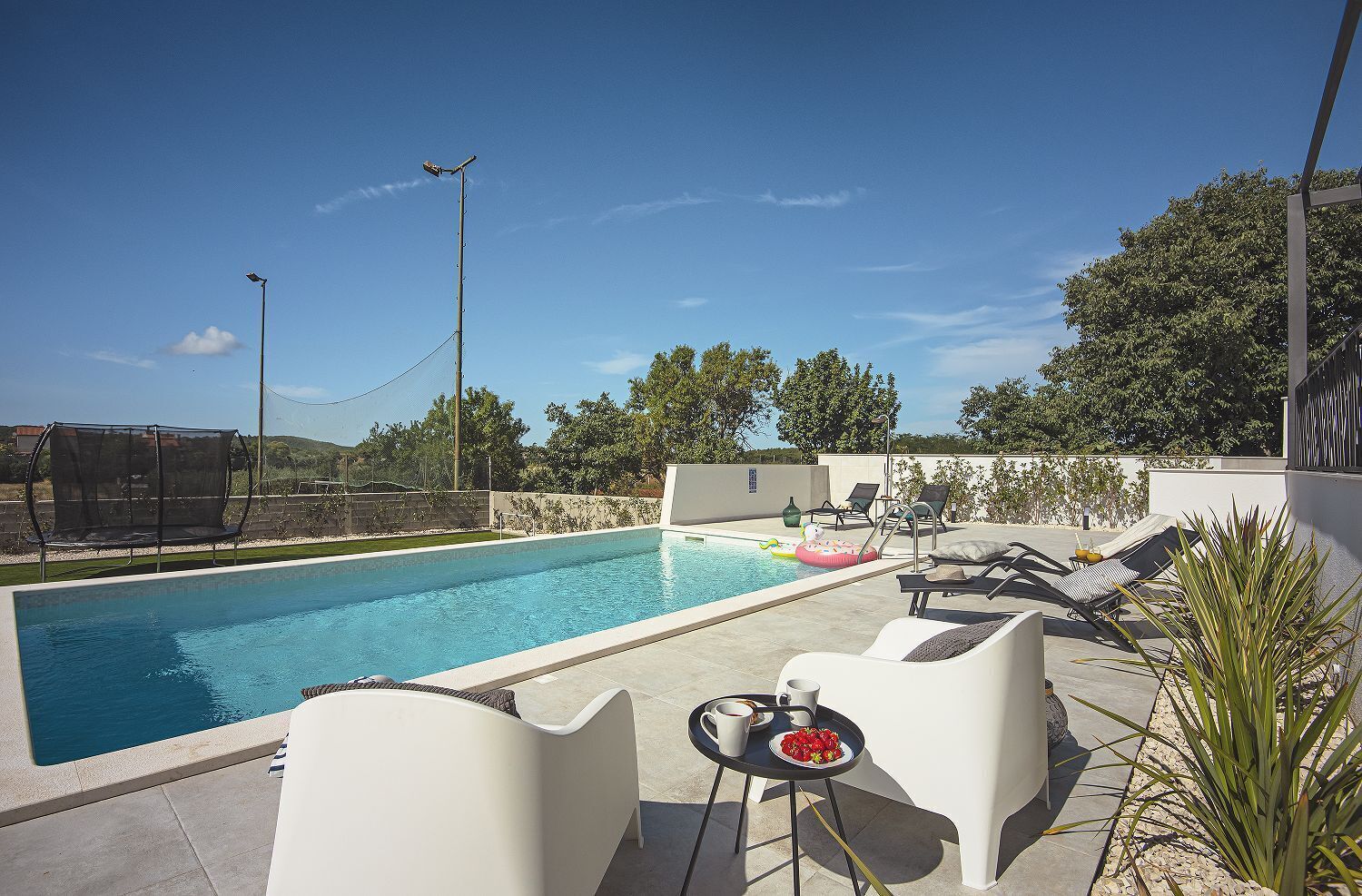 En swimmingpool med liggestole og et bord, omgivet af en stenvæg og grøn vegetation.