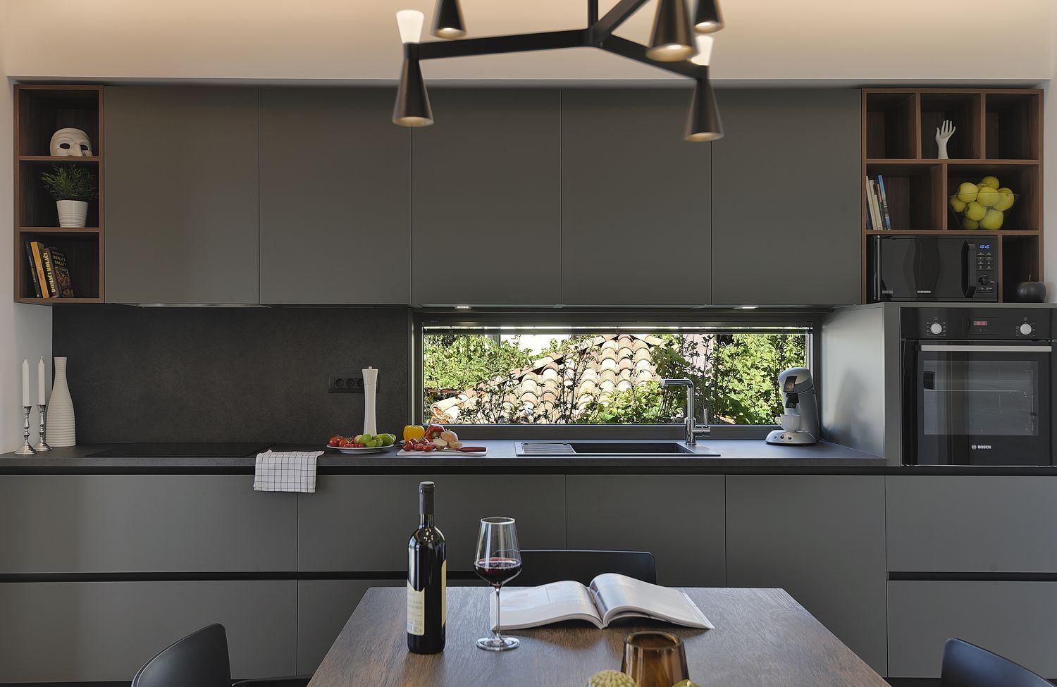 Une cuisine moderne dotée d’un four encastré, d’une fenêtre donnant sur un espace verdoyant et d’un coin repas avec une table et des chaises.