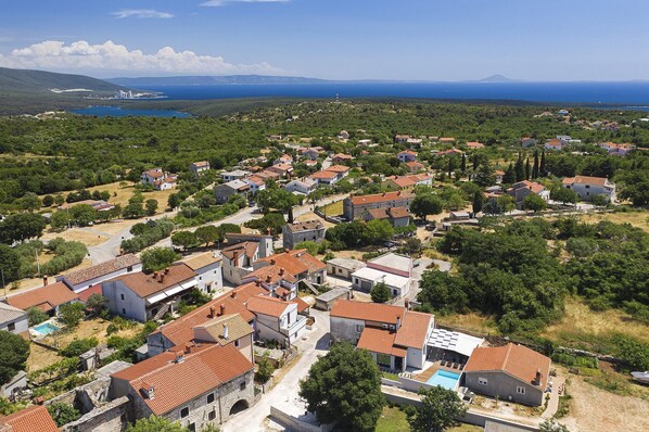 Vue aérienne d’un village côtier doté d’une architecture traditionnelle, avec une place centrale et une vue sur la mer et les montagnes au loin.