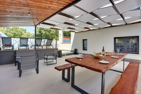An outdoor patio with a wooden table set for dining, surrounded by chairs, and a pool in the background.