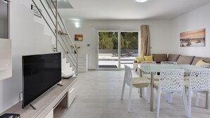 Ein modernes Wohnzimmer mit Flachbildfernseher, einem Essbereich mit Glastisch und weißen Stühlen, einer Couch mit Kissen und einer Treppe.