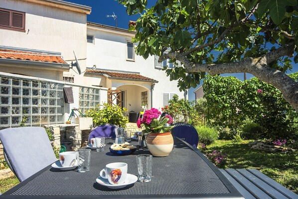 Un área de comedor al aire libre con una mesa para dos, rodeada de follaje y un edificio al fondo.