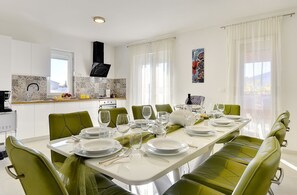 A dining area with a long table set for six, green chairs, and a kitchen in the background.