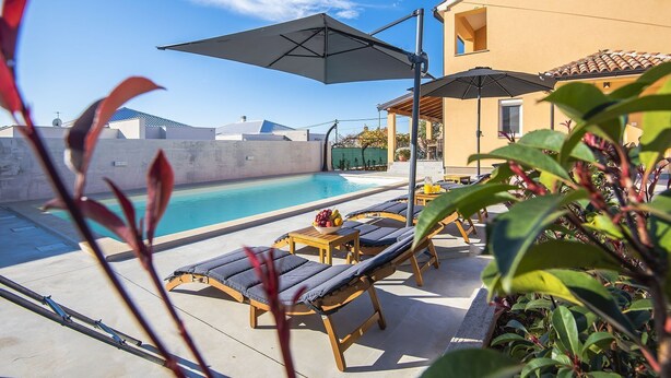 A poolside area with lounge chairs and umbrellas, a clear blue sky, and a building in the background. - Villa with private pool, playground, WiFi, air conditioning, BBQ (Pula)