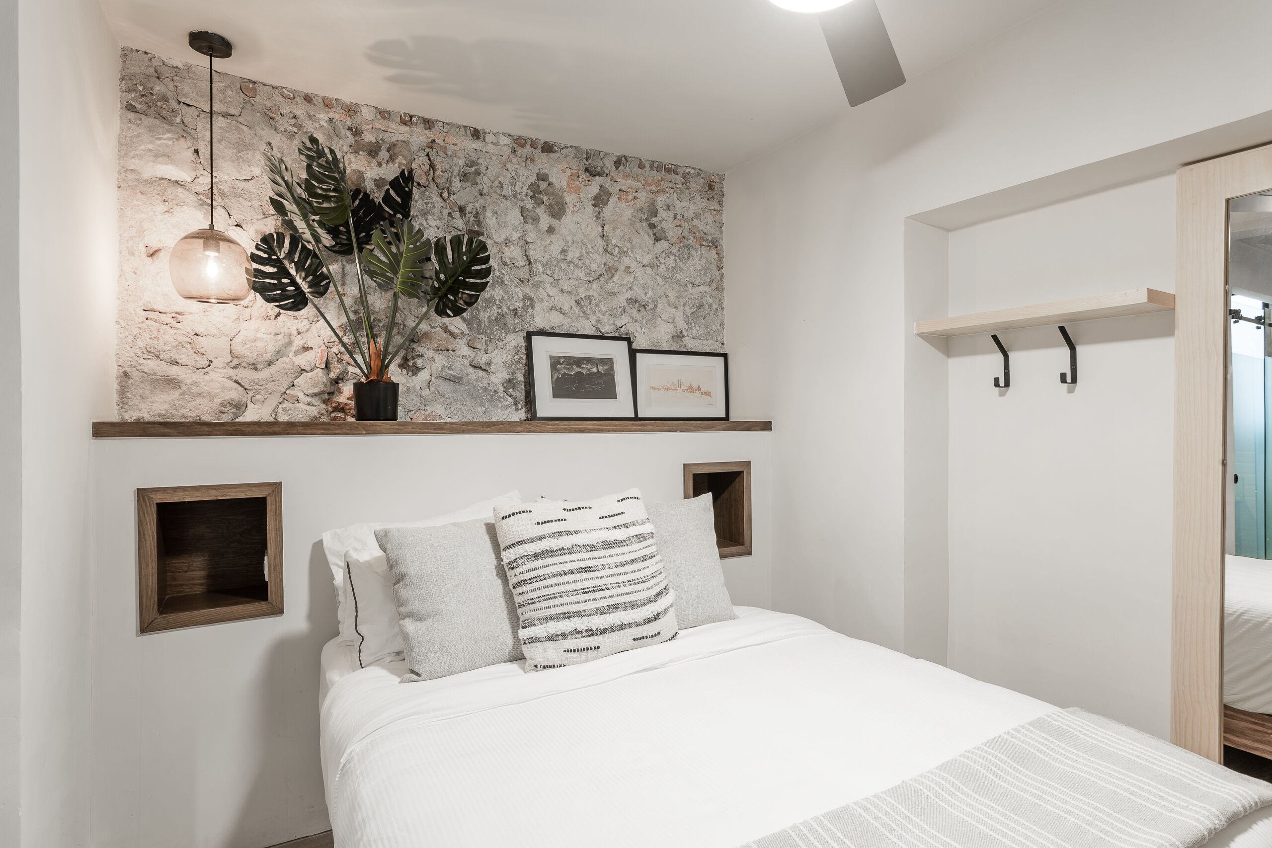 Un dormitorio con pared de piedra, una lámpara colgante, una planta en maceta, cuadros enmarcados y una cama con almohadas.