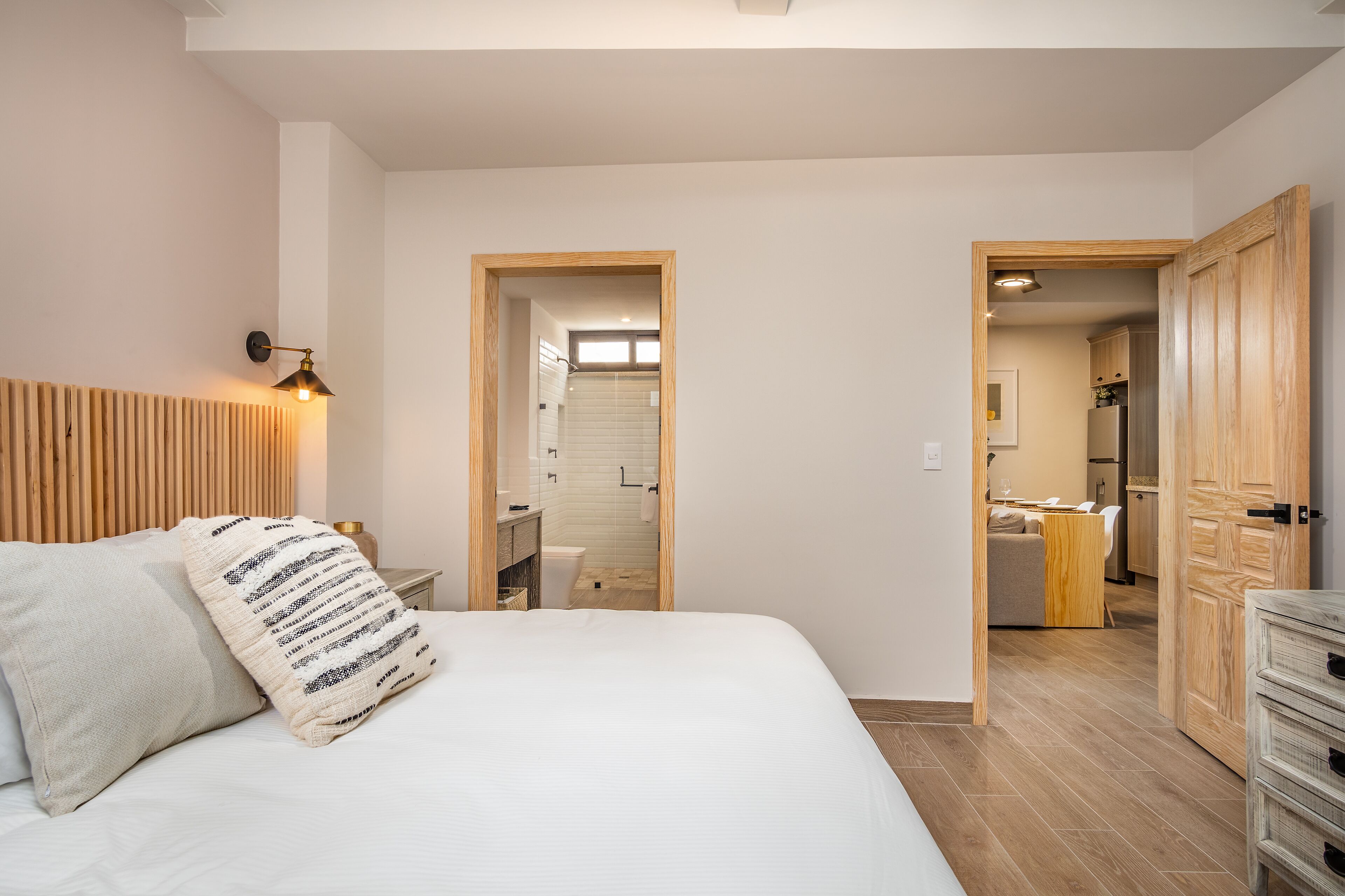 A bedroom with a bed, a bedside lamp, a wooden headboard, and a view into the bathroom and kitchen through an open door.