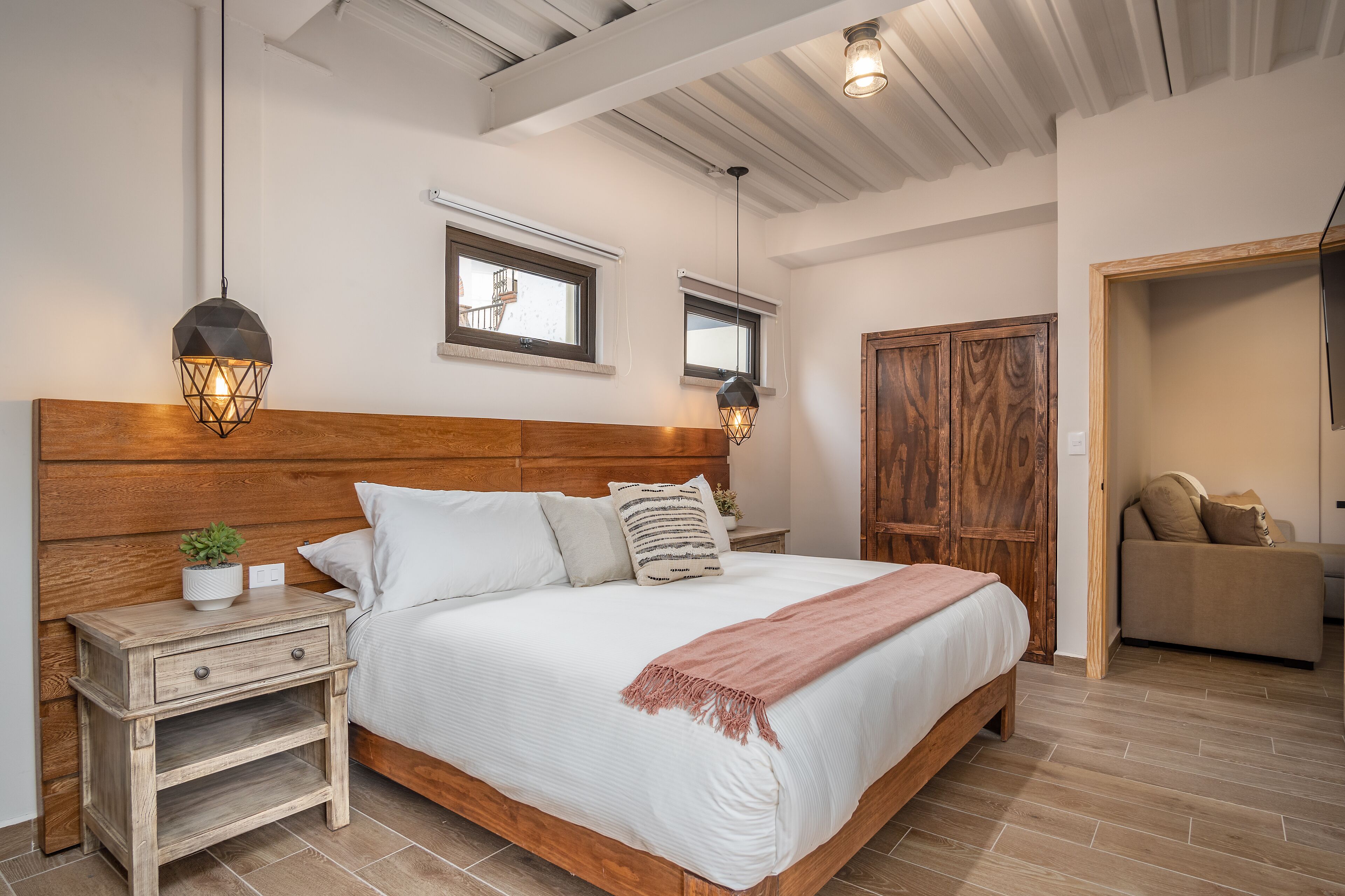 A bedroom with a wooden headboard, a bedside table with a drawer, a bedside lamp, a window, and a door leading to another room.
