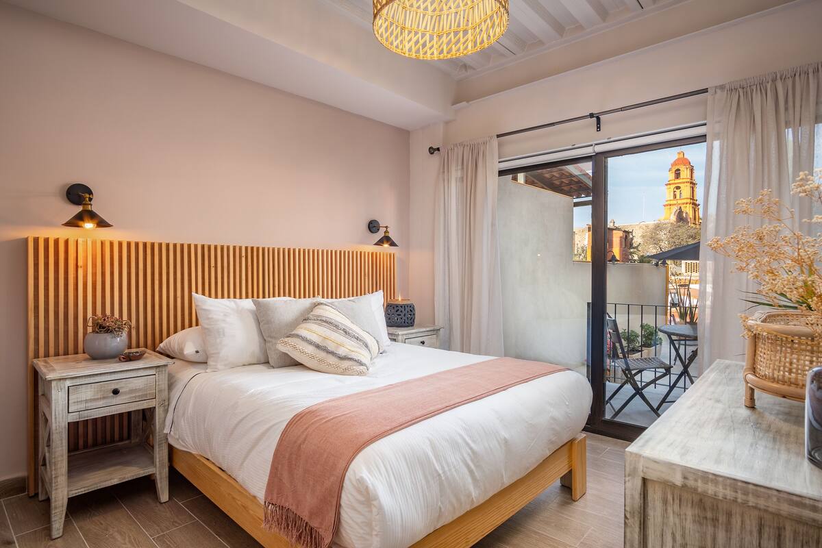 Een slaapkamer met een houten hoofdeinde, een groot bed met een roze plaid, twee nachtkastjes met lampen, een uitzicht op een gebouw door een glazen schuifdeur, en een balkon met tuinmeubilair.
