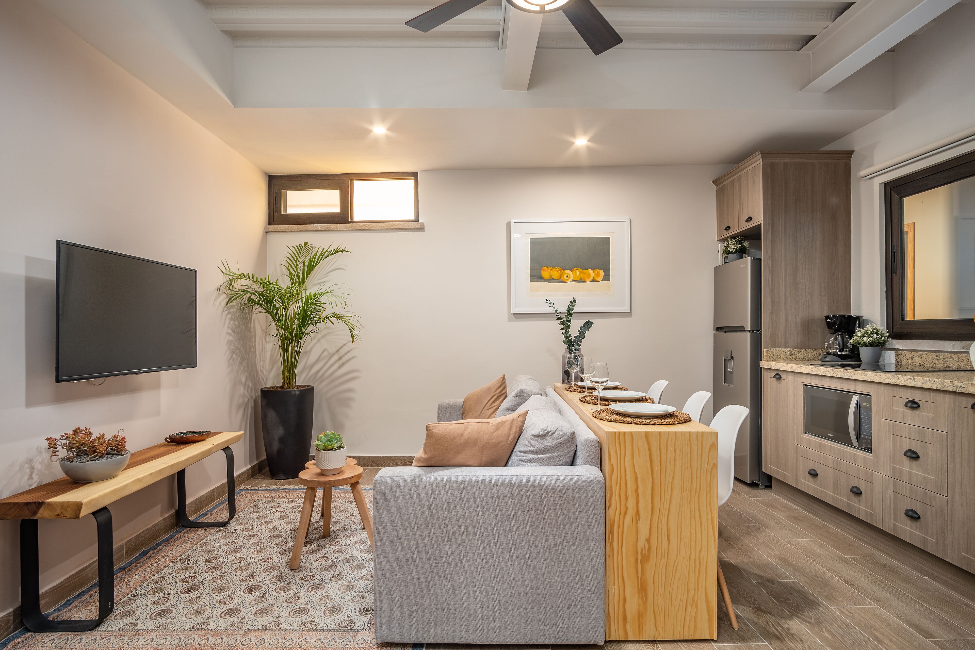 A modern living room with a grey sofa, wooden coffee table, and a flat-screen TV. A small round table with a potted plant is in the corner. The kitchen area is visible with wooden cabinets and a built-in oven.