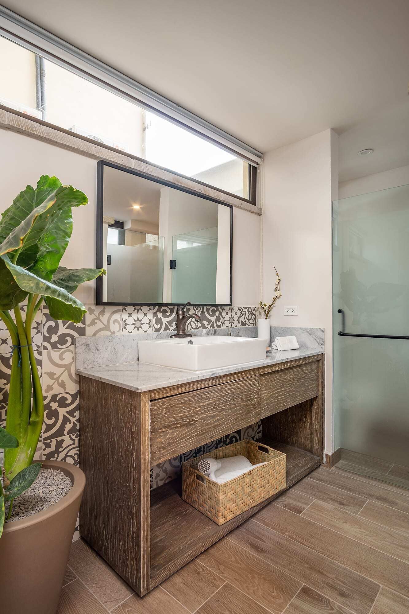 A bathroom with a large mirror, a white sink, a wooden vanity, a potted plant, and a woven basket.