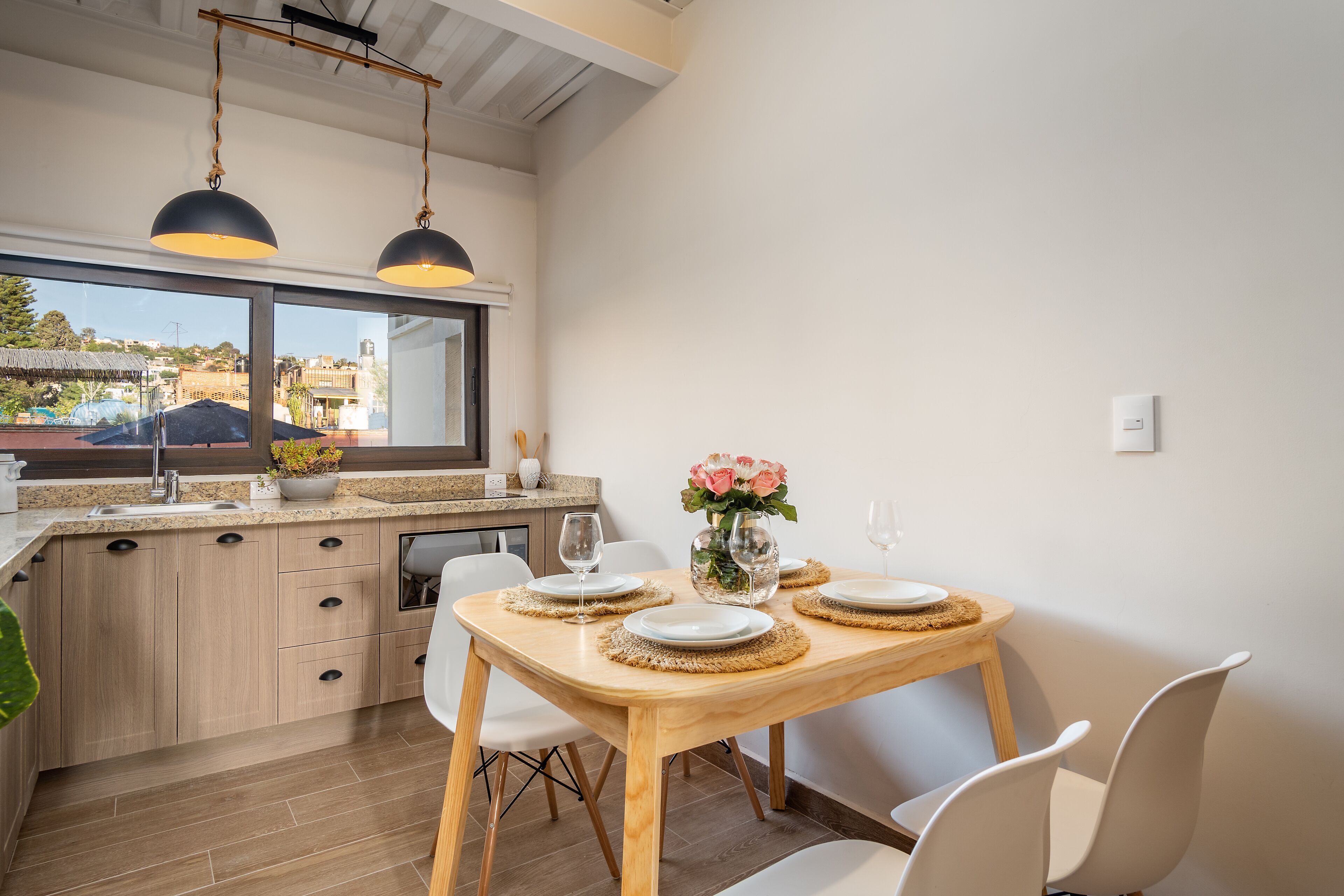 A modern kitchen with wooden cabinets, a dining table set with plates and a vase of flowers, and a large window offering a view of the outdoors.