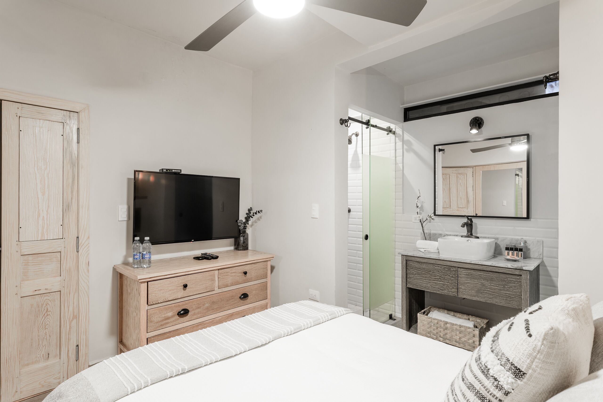 A bedroom with a bed, wooden dresser, TV, and a bathroom with a mirror and sink.