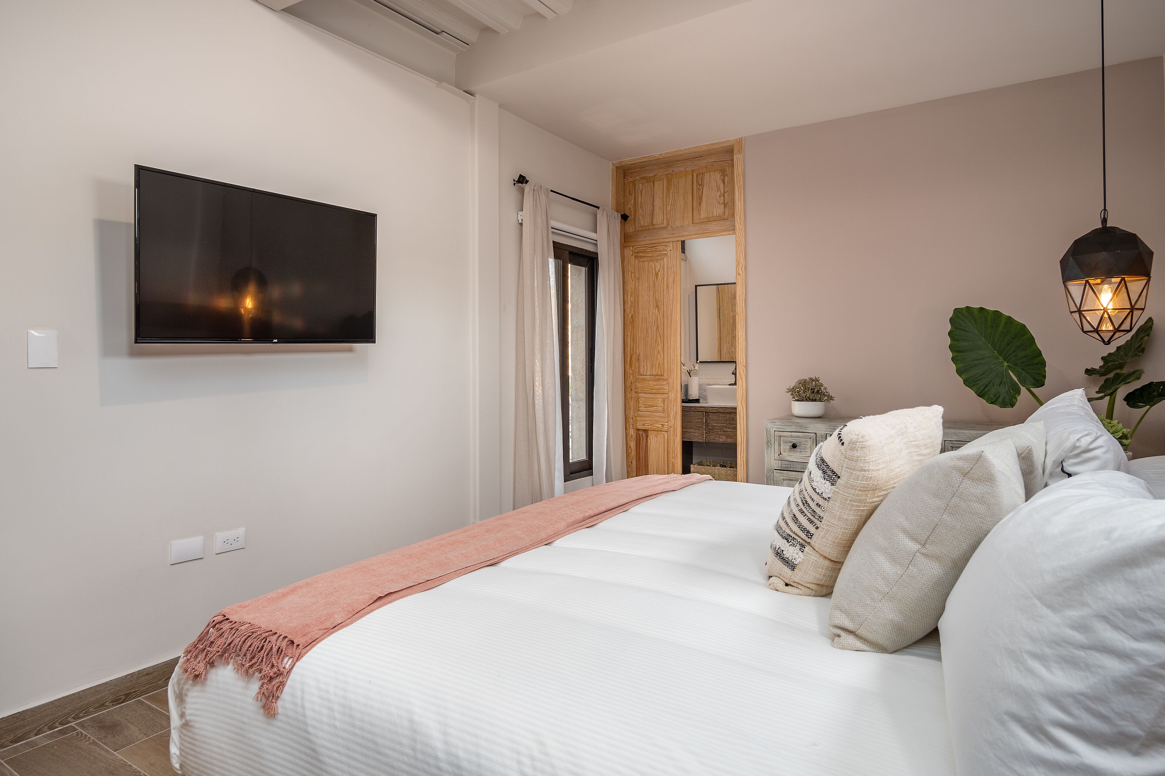 A bedroom with a bed, a television mounted on the wall, a wooden door, and a hanging light fixture.
