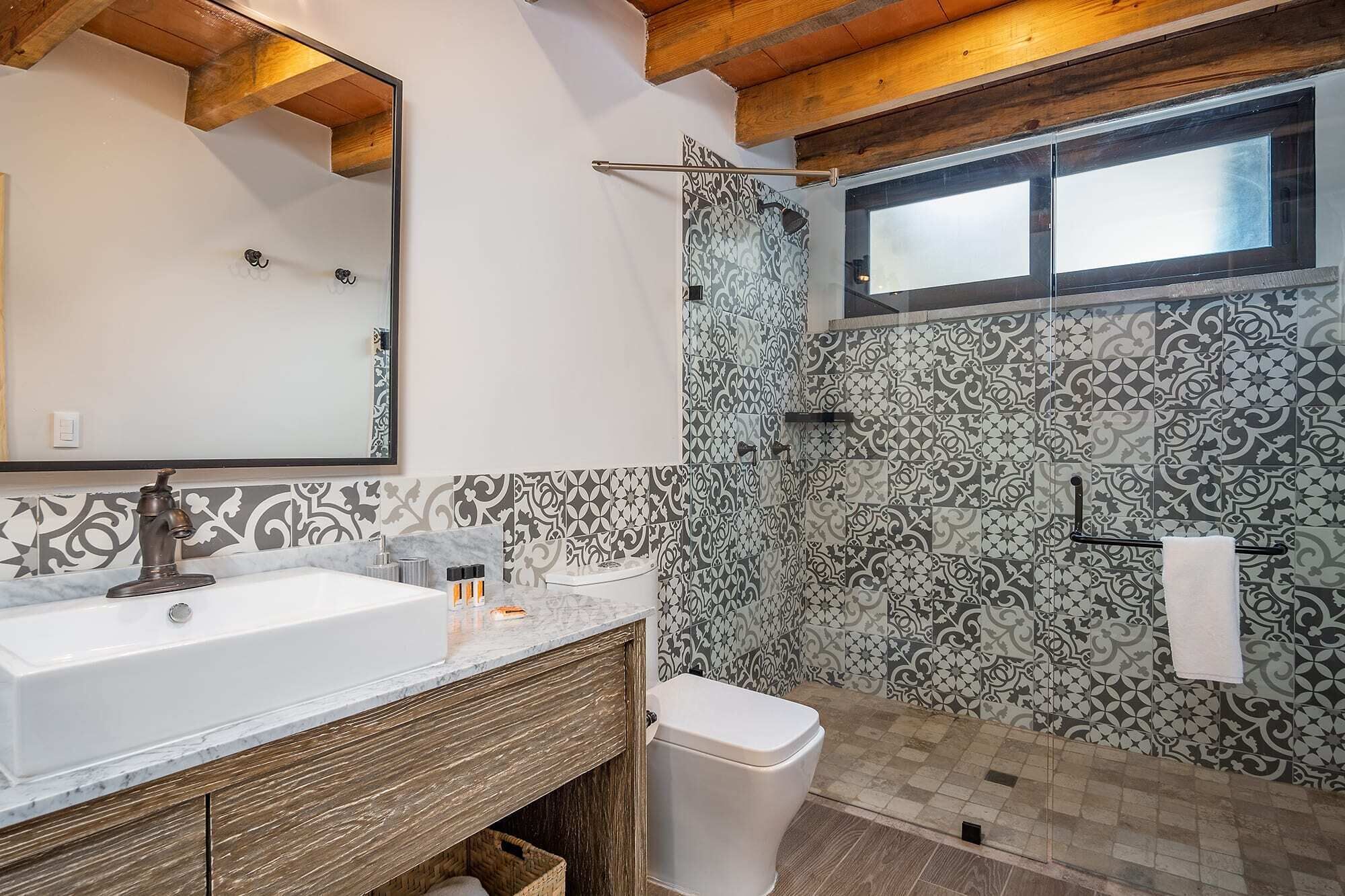 A bathroom with a white sink, wooden vanity, patterned tiles, a shower, and a window.