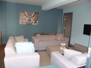 A living room with a beige sofa, a white armchair, and a small coffee table. There is a painting of an owl on the wall and a door in the background. - Brabander Alm (Biberwier)