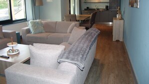 Una sala moderna con un sofá, un sillón y una mesita con un control remoto. Se ve una cocina al fondo con una mesa de comedor y sillas.